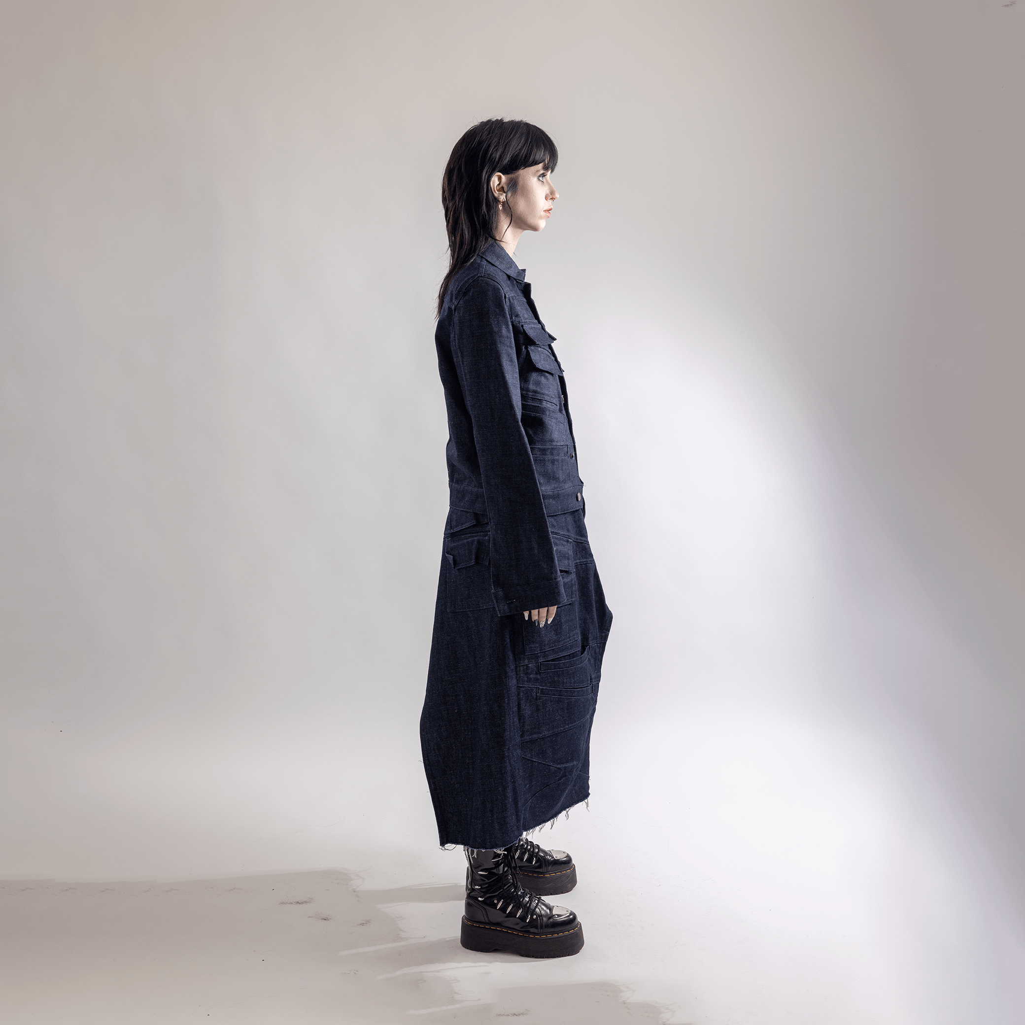 Woman in a long dark denim coat and boots standing sideways in a minimalist studio setting